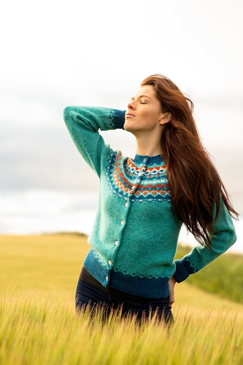 Women standing in a field of wheat wearing an Aqua colour wool fair isle cardigan. Crathie pattern with orange and teal.