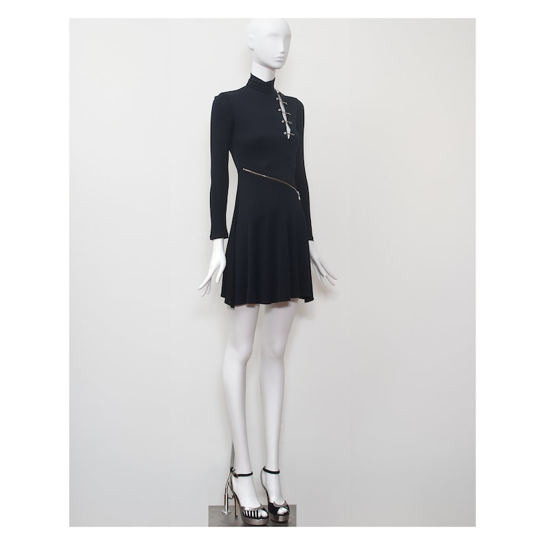 May include: A black mini dress with a high neckline and long sleeves. The dress has a zipper detail on the side and a row of safety pins down the front.
