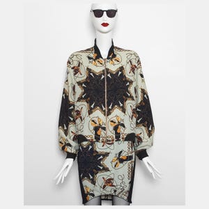 Könnte beinhalten: Ein zweiteiliges Outfit auf einer Schaufensterpuppe. Das Set beinhaltet eine Bomberjacke und einen Rock. Die Jacke und der Rock sind hellgrün mit einem schwarzen und goldenen Sternmuster. Die Jacke hat einen schwarzen Kragen, Manschetten und einen Reißverschluss.
