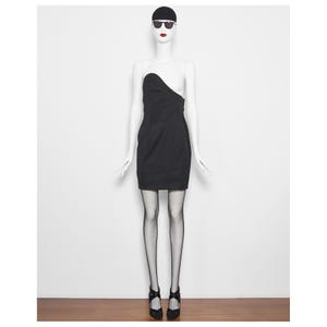 May include: A black, one-shoulder mini dress on a mannequin. The dress has a fitted silhouette and an asymmetrical neckline. The mannequin is wearing black fishnet stockings and black heels. The mannequin is also wearing a black cap and sunglasses.