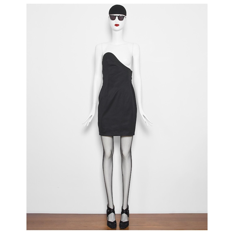 May include: A black, one-shoulder mini dress on a mannequin. The dress has a fitted silhouette and an asymmetrical neckline. The mannequin is wearing black fishnet stockings and black heels. The mannequin is also wearing a black cap and sunglasses.