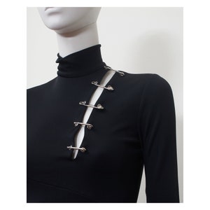 May include: Black long-sleeved shirt with a mock neck and a diagonal cut-out detail on the front. The cut-out is secured with silver safety pins.