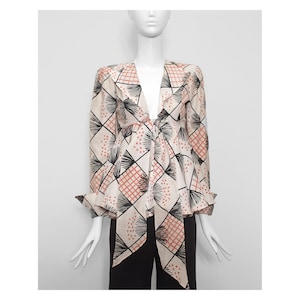 Puede incluir: Una blusa blanca con un patrón geométrico en negro, naranja y rosa. La blusa tiene un escote en V profundo y mangas largas con un lazo en la cintura.