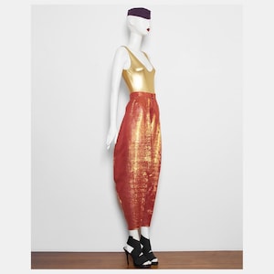 May include: A mannequin wearing a gold metallic bodysuit and high-waisted, red and gold patterned trousers. The mannequin also wears a purple hat and black and white high-heeled sandals. The outfit is displayed against a white background.