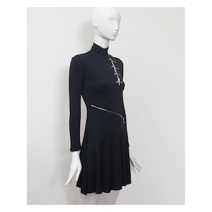 May include: A black mini dress with long sleeves and a high neckline. The dress has a diagonal zipper detail and a row of safety pins down the front.