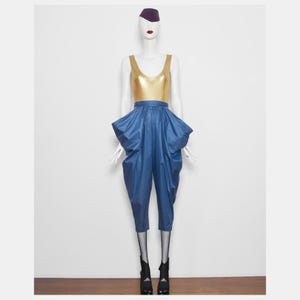 Puede incluir: Un maniquí exhibe una camiseta sin mangas dorada metálica combinada con pantalones azules de cintura alta y fruncidos. El atuendo se completa con un sombrero morado, medias de rejilla negras y botines negros. El maniquí tiene pintalabios rojo.