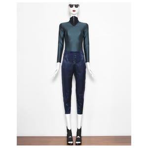 Puede incluir: Un maniquí exhibe una parte superior de manga larga color verde azulado con cuello negro y cremallera, combinada con pantalones azul marino de cintura alta. Los pantalones tienen cierre de botones. El maniquí lleva sandalias de tacón negras y blancas y gafas de sol.