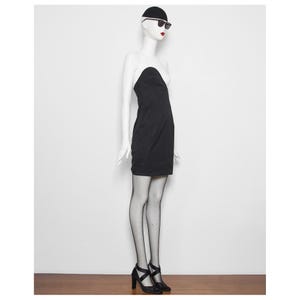 May include: A black strapless dress on a mannequin. The dress is worn with black heels, fishnet stockings, and sunglasses. The mannequin is wearing a black cap. The background is a plain white wall and a wooden floor.