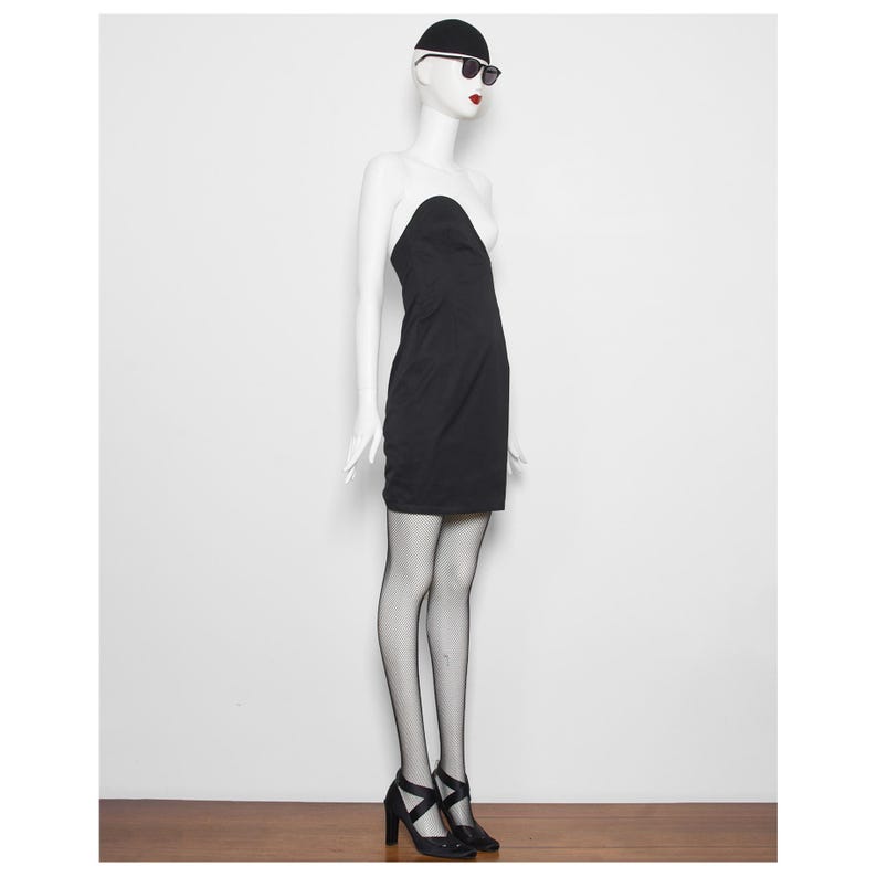 May include: A black strapless dress on a mannequin. The dress is worn with black heels, fishnet stockings, and sunglasses. The mannequin is wearing a black cap. The background is a plain white wall and a wooden floor.