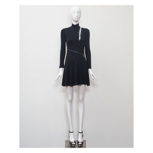 May include: A black mini dress with long sleeves and a high neckline. The dress has a diagonal zipper and a row of silver safety pins on the front.