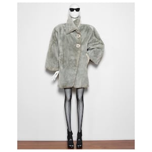 Puede incluir: Un abrigo de piel sintética gris con cuello y dos botones. El abrigo está en un maniquí que lleva medias negras de lunares blancos y zapatos negros.