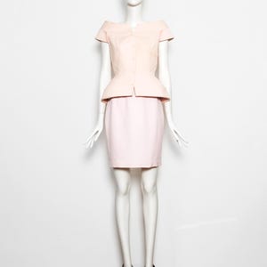 May include: A light pink two-piece suit displayed on a mannequin. The suit includes a short-sleeved, button-down top with a peplum waist and a matching pencil skirt. The mannequin is wearing black sunglasses and black heels.