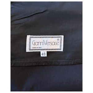 May include: A close-up of a black garment with a white label. The label reads "Gianni Versace MADE IN ITALY" and features the number "40". The label is rectangular with a decorative border.