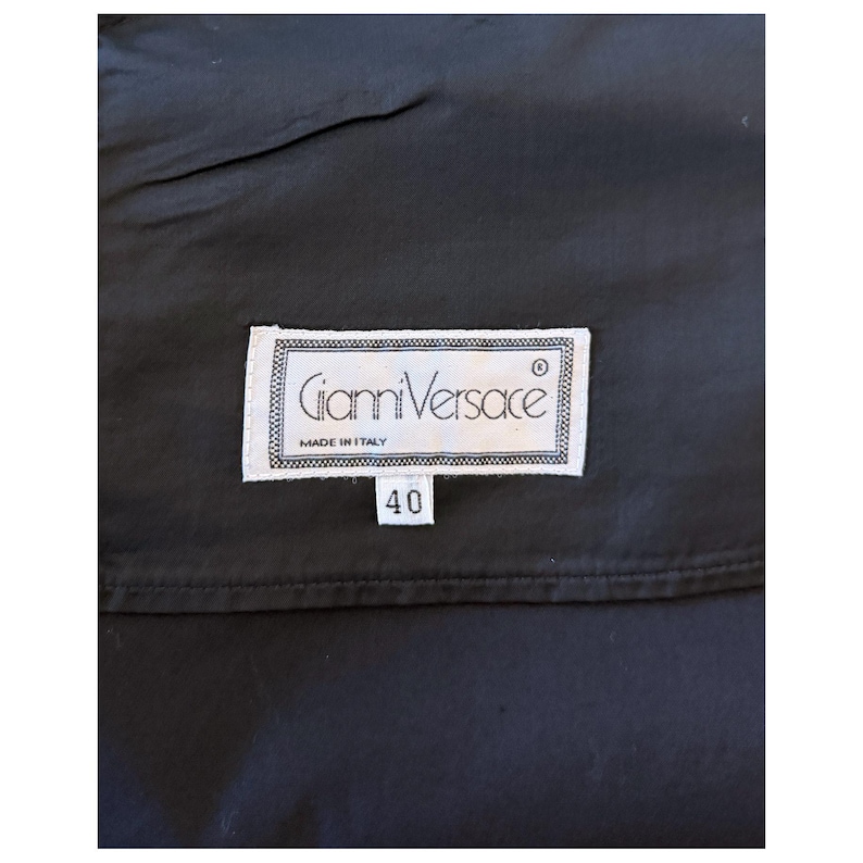 May include: A close-up of a black garment with a white label. The label reads "Gianni Versace MADE IN ITALY" and features the number "40". The label is rectangular with a decorative border.
