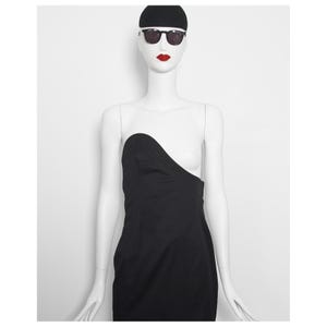 May include: A black strapless dress on a mannequin. The dress features an unusual asymmetrical design. The mannequin is wearing sunglasses, a black cap, and red lipstick. The background is plain white.