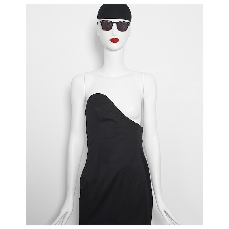 May include: A black strapless dress on a mannequin. The dress features an unusual asymmetrical design. The mannequin is wearing sunglasses, a black cap, and red lipstick. The background is plain white.