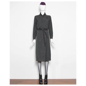 Könnte beinhalten: Ein graues und schwarzes gestreiftes Kleid mit einem Mandarin-Kragen und einem schwarzen geflochtenen Gürtel. Das Kleid ist knielang und hat lange Ärmel. Das Model trägt schwarze Strumpfhosen mit weißen Punkten und schwarze Schuhe mit Absatz.