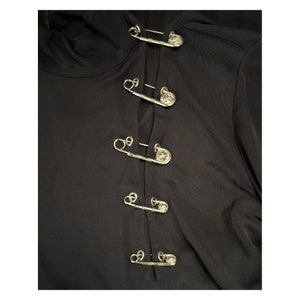May include: Black long-sleeved shirt with a row of silver safety pins with decorative heads down the front.