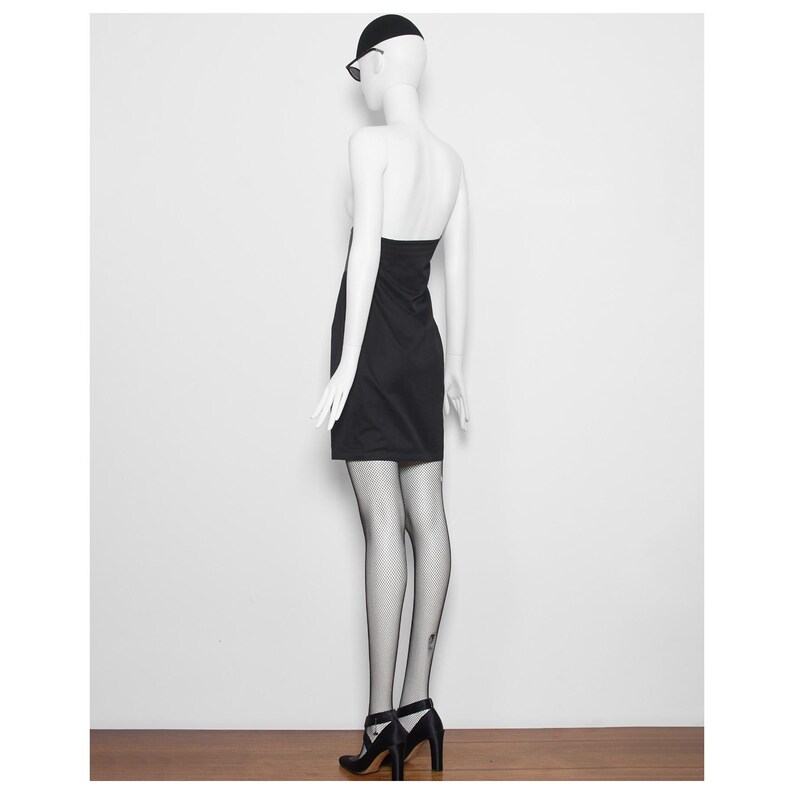 May include: A black halter dress on a mannequin. The dress is knee-length and paired with black fishnet stockings and black heels. The mannequin is also wearing a black cap and sunglasses.