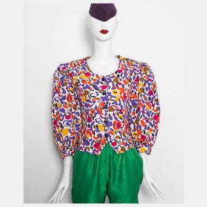 May include: A patterned blouse in a variety of colours, including purple, orange, and yellow. The blouse has a scoop neckline and long sleeves. The outfit is completed with bright green trousers. A purple hat is on the mannequin's head.