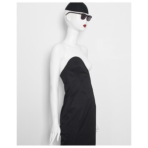May include: A black strapless dress on a mannequin. The mannequin is wearing a black cap and sunglasses. The mannequin has red lipstick. The dress is a simple, elegant design.