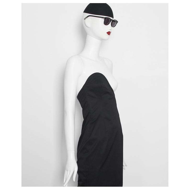 May include: A black strapless dress on a mannequin. The mannequin is wearing a black cap and sunglasses. The mannequin has red lipstick. The dress is a simple, elegant design.