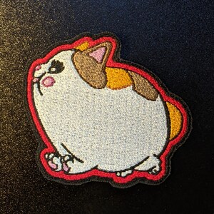 May include: A white, brown, and orange embroidered patch of a cartoon cat with red outlines. The cat is looking up and has a round, chubby body.