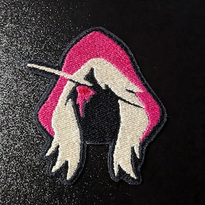 May include: A pink hooded figure with long white hair, a single red drop of blood on the forehead, and a white stick protruding from the head. The figure is embroidered on a black fabric.
