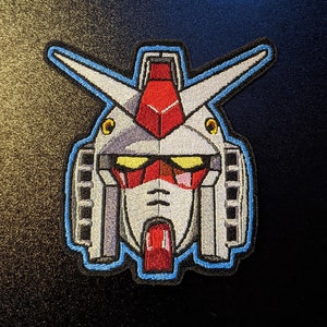 May include: Embroidered patch of a white and gray robot head with red and yellow accents. The robot has a blue outline and is set against a dark background.
