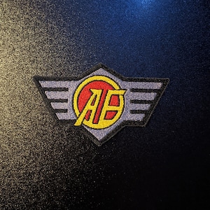 Op de afbeelding: Een geborduurde patch in zwart en wit met een rode en gele cirkel met de letters "AB" en vleugels aan beide kanten.