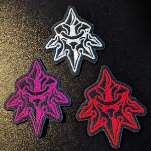 Op de afbeelding: Drie geborduurde patches in de vorm van een gestileerde ster met een gezicht in het midden. De patches zijn wit, roze en rood.