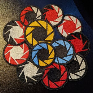 May include: A collection of ten embroidered patches featuring the Aperture Science logo in various colors, including red, yellow, blue, black, and white.