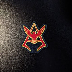May include: A red and yellow embroidered patch with a stylized eagle design inside a triangle shape.