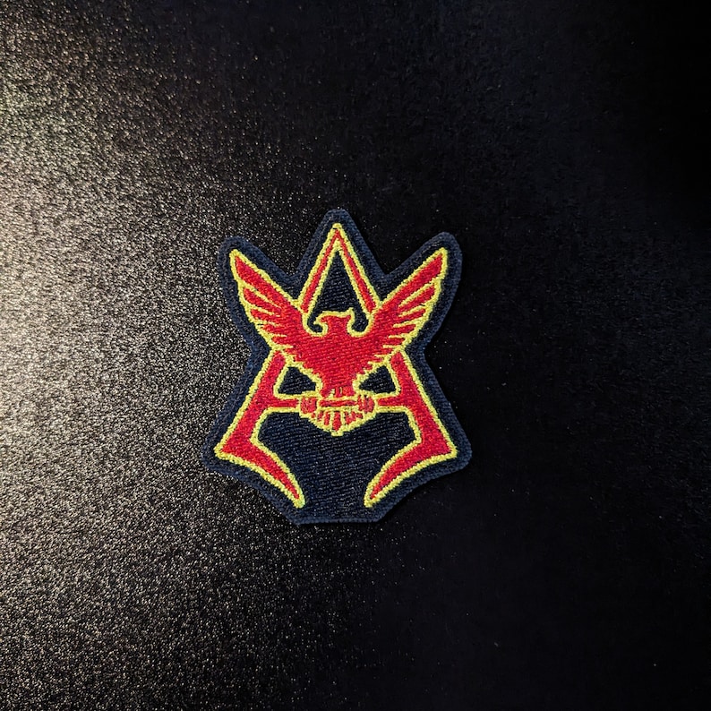 May include: A red and yellow embroidered patch with a stylized eagle design inside a triangle shape.