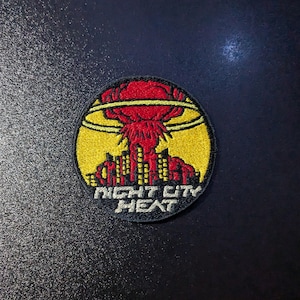 May include: A black and yellow embroidered patch with a red mushroom cloud over a city skyline. The text "Night City Heat" is written in a black banner below the image.