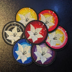 May include: Seven embroidered patches featuring a white flower with a yellow center on a variety of colored backgrounds. The patches are circular and have a black border.