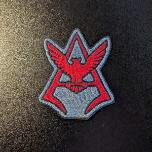 May include: A red and blue embroidered patch with a stylized eagle design. The eagle is centered in a triangle shape with a red outline.