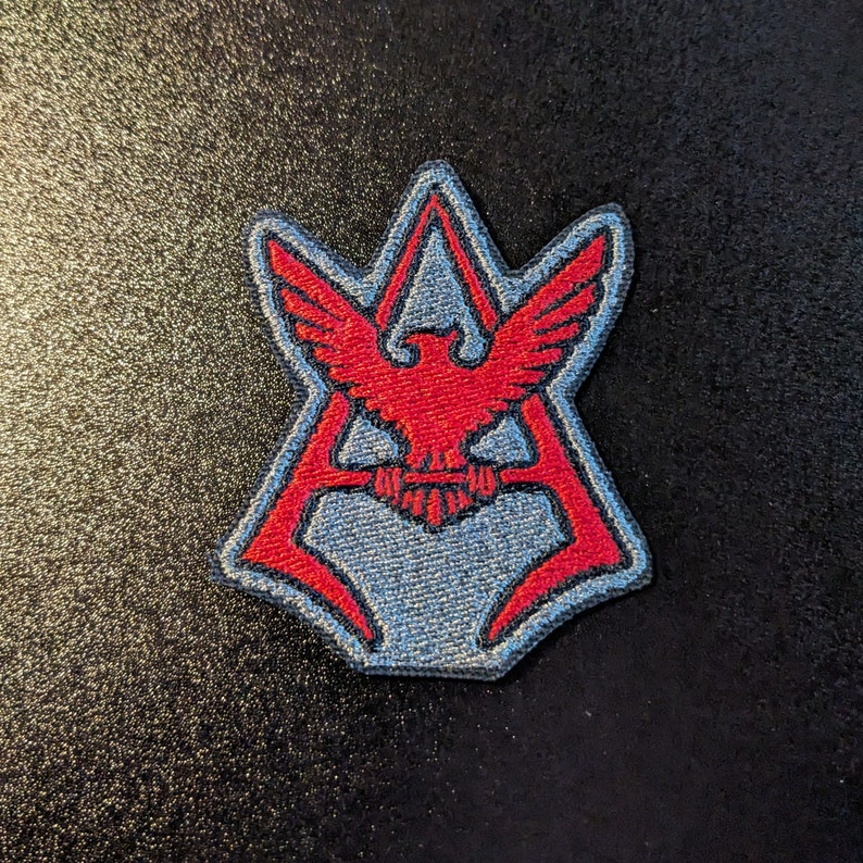 May include: A red and blue embroidered patch with a stylized eagle design. The eagle is centered in a triangle shape with a red outline.