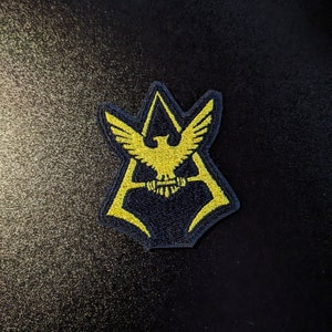 May include: A yellow embroidered patch with a stylized eagle design inside a black triangle shape.