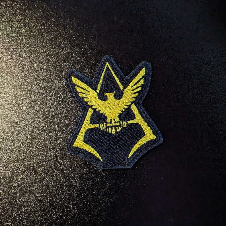 May include: A yellow embroidered patch with a stylized eagle design inside a black triangle shape.