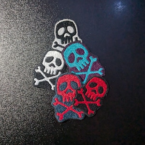 May include: Five embroidered skull and crossbones patches in different colors. The patches are arranged in a pile, with the white skull on top.