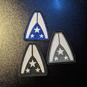 May include: Three embroidered patches in the shape of a triangle with a white star design. The patches are blue, gray, and black.