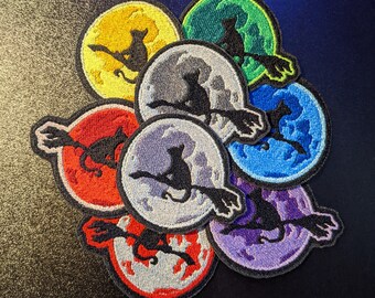 May include: Eight embroidered patches featuring black cats riding broomsticks on different colored moons. The moons are yellow, green, gray, blue, purple, red, and two white moons.