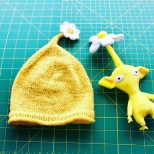 Könnte beinhalten: Eine gelbe Strickmütze mit einer weißen Blume oben, neben einem gelben Plüschtier mit einer weißen Blume auf dem Kopf.