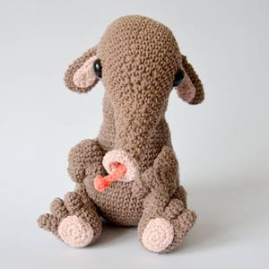 Può includere: Un peluche marrone lavorato a uncinetto con orecchie e piedi rosa. L'animale ha un lungo naso marrone e occhi neri. È seduto su una superficie bianca.