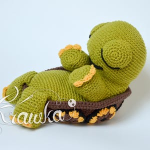 Pode incluir: Uma tartaruga de crochê verde com os olhos fechados, deitada de costas dentro de uma concha de crochê marrom e amarela.