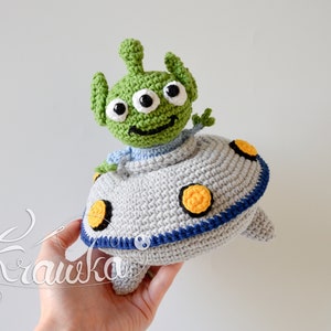 Op de afbeelding: Een gehaakte groene alien met drie ogen en een glimlachend gezicht zit in een grijze gehaakte vliegende schotel met gele en blauwe accenten.