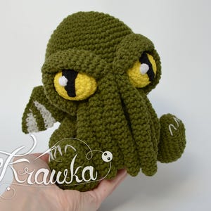 May include: A handmade, olive green crochet Cthulhu plushie with large yellow and black eyes. The tentacles and wings are detailed with white accents. The plushie is held in a hand, with the artist's signature "Krawka" in cursive.