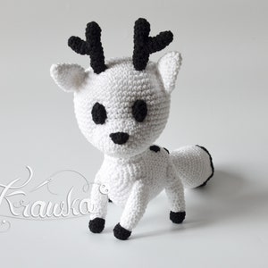 May include: White crocheted deer with black antlers, eyes, nose, and feet. The deer is standing on a white surface.