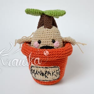 Può includere: Un giocattolo amigurumi all'uncinetto di una pianta di mandragola in un vaso di terracotta. La pianta ha uno stelo marrone, foglie verdi e un viso beige con guance rosa. Il vaso ha la scritta "MANDRAKE".